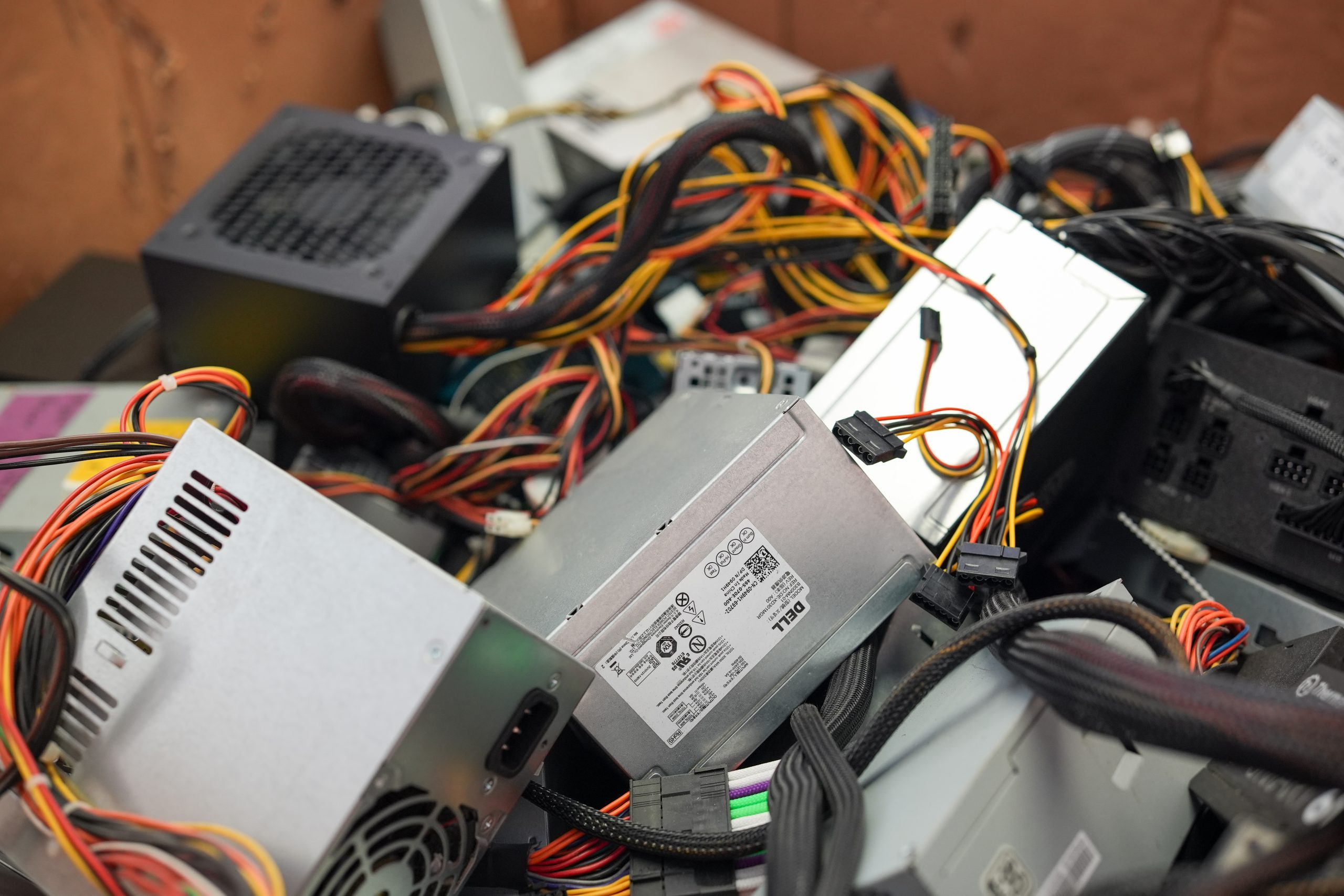 A pile of various computer power supply units with tangled cables and connectors, destined for electronics recycling, rests in a cardboard box. Some units sport labels and logos, with wires in red, yellow, and black contrasting sharply beneath the Omega ECycles banner committed to data destruction.