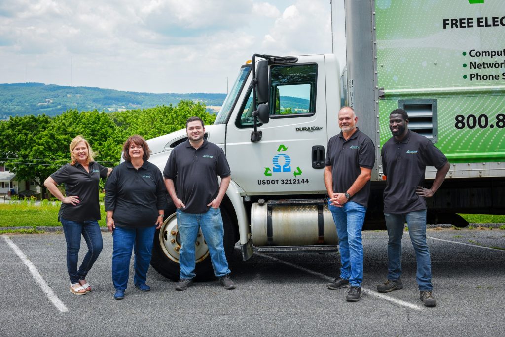 Five members of the Omega ECycles team, wearing matching black shirts, stand smiling in front of a white and green company truck on a sunny day, with trees and hills in the background.