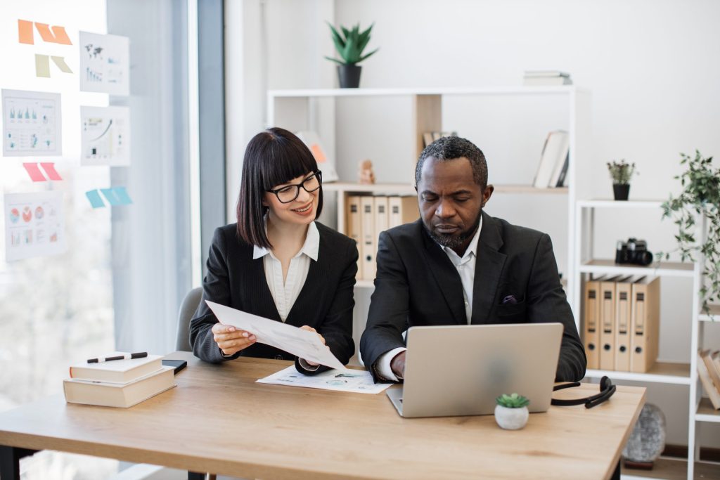 Two business professionals sit at a desk in a Central PA office. The woman smiles while holding documents, and the man works on a laptop, possibly discussing data destruction of old devices. Shelves, books, and plants add warmth to the workspace.