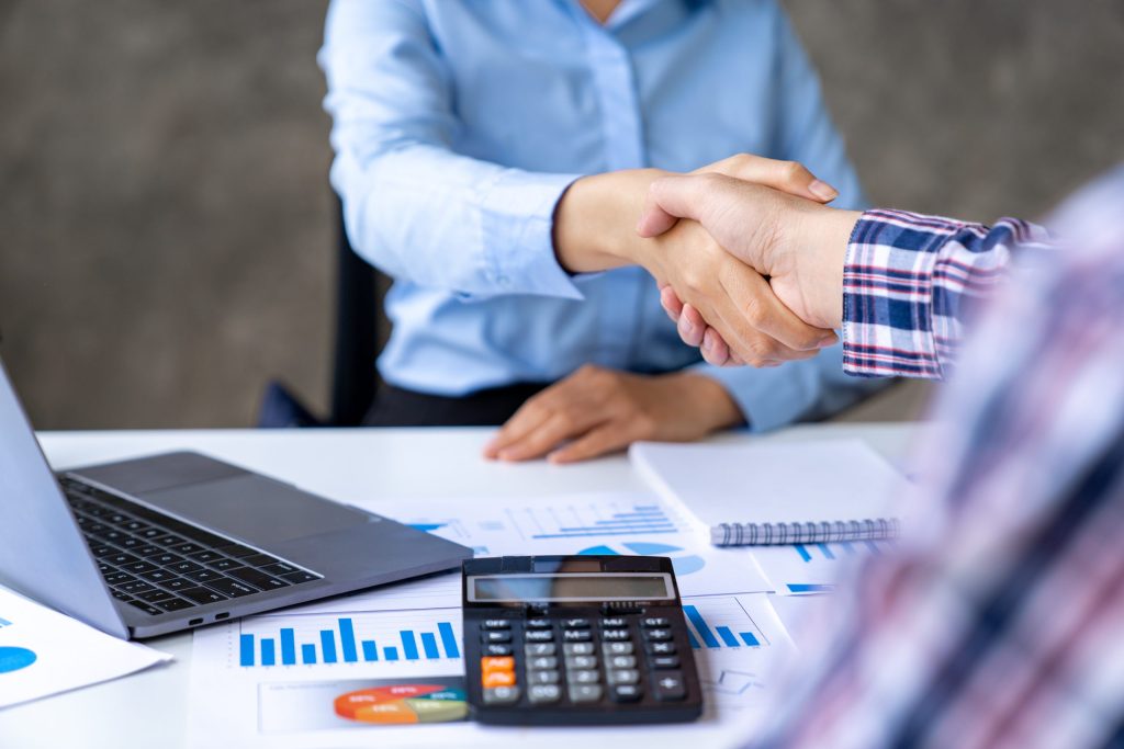 Two people shaking hands over a desk with documents, charts, and a laptop, suggesting a successful Omega ECycles business meeting focused on electronics recycling and giving old devices a new purpose.