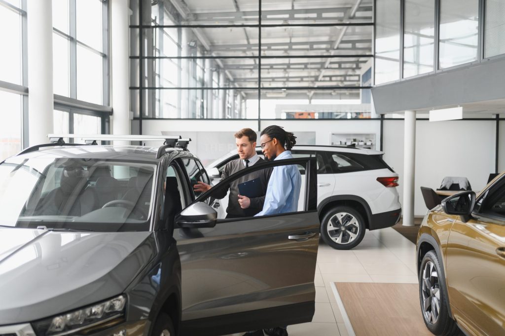 Two men stand beside a gray SUV inside a bright car dealership showroom, with one holding a clipboard and both looking at the car's interior. The dealership also offers electronics recycling for car dealerships, ensuring secure data destruction. Other vehicles are visible in the background.