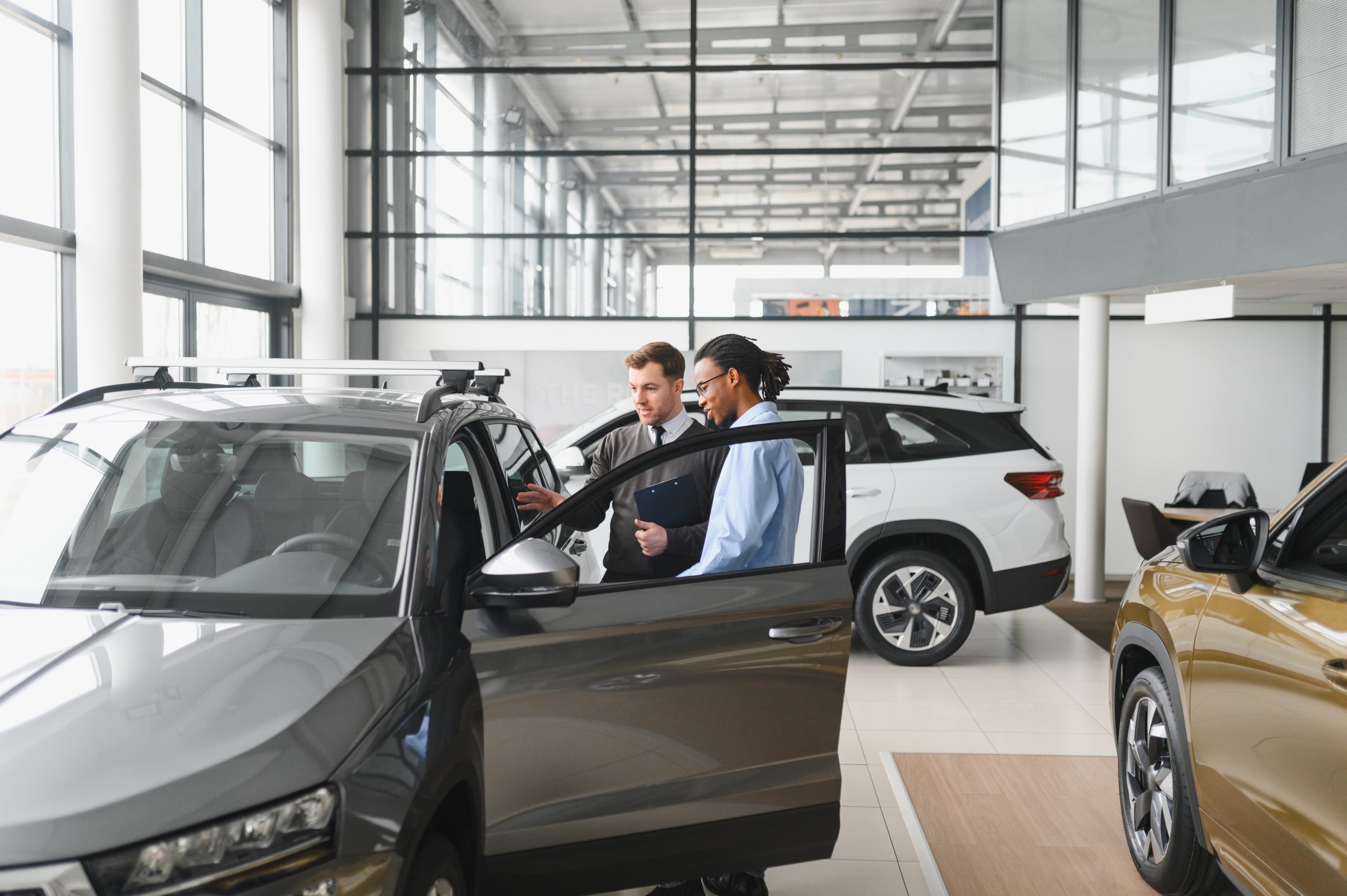 Two men stand beside a gray SUV inside a bright car dealership showroom, with one holding a clipboard and both looking at the car's interior. The dealership also offers electronics recycling for car dealerships, ensuring secure data destruction. Other vehicles are visible in the background.