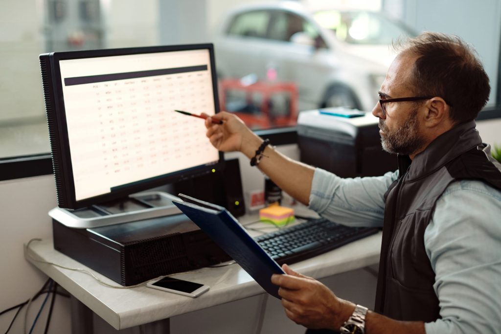 A man wearing glasses sits at a desk, pointing at data on a computer monitor with a pen while holding a clipboard. Electronics recycling for car dealerships and data destruction tasks are visible on the screen. A car is outside the window in the background.