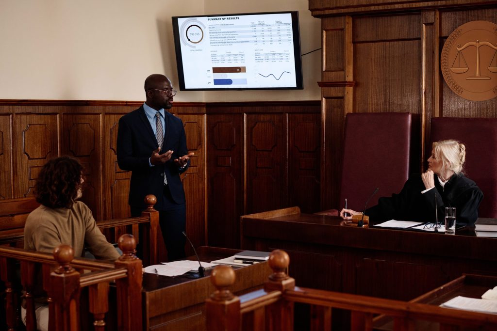 A man in a suit presents data on a screen in a courtroom, addressing the repercussions of improper data handling to a judge and others, highlighting the importance of services like Omega ECycles and secure data destruction for law firms.