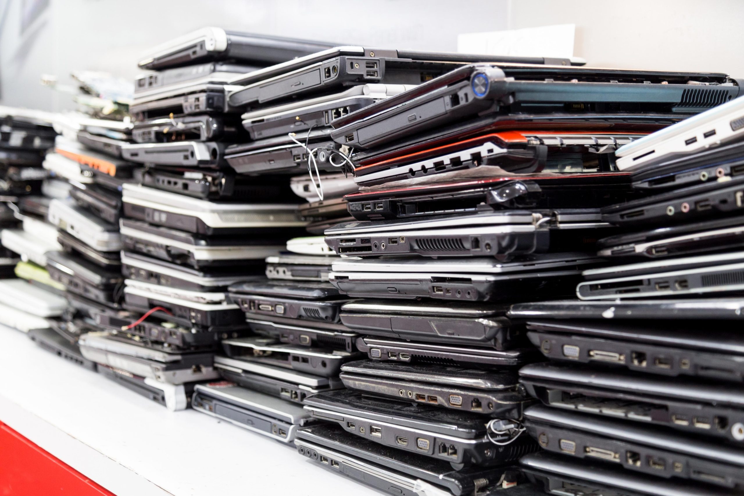 Stacks of old, used laptops piled on a white surface, showing various models and visible wear—possibly collected for an electronics recycling or computer recycling initiative.