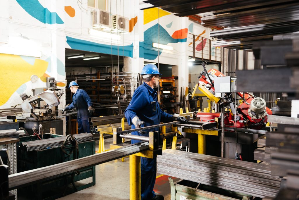 Workers in blue uniforms and helmets operate machinery and handle metal bars in a brightly lit industrial workshop focused on electronics recycling for manufacturers, with colorful wall murals and neatly organized equipment.