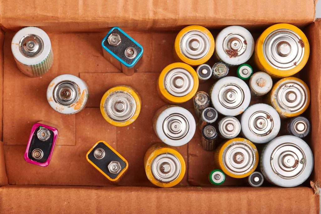 A cardboard box containing various used batteries, including AA, AAA, C, D, and rectangular 9V types—all ready for recycling old batteries—seen from above with some showing signs of rust and wear.
