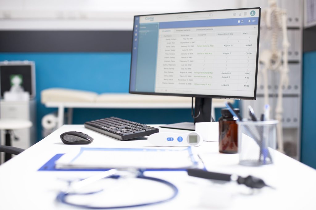 A desk in a medical office with a computer displaying patient appointments, keyboard, mouse, clipboard, pen holder, medicine bottle, and a stethoscope; HIPAA-compliant data destruction ensures sensitive info is safe when electronics recycling for healthcare.