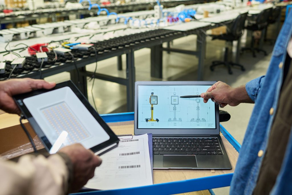 Two people stand at a workbench in a factory, one holding an LED panel, the other pointing at a technical diagram on a laptop. Rows of electronics and workstations dedicated to electronics recycling for manufacturers are visible in the background.