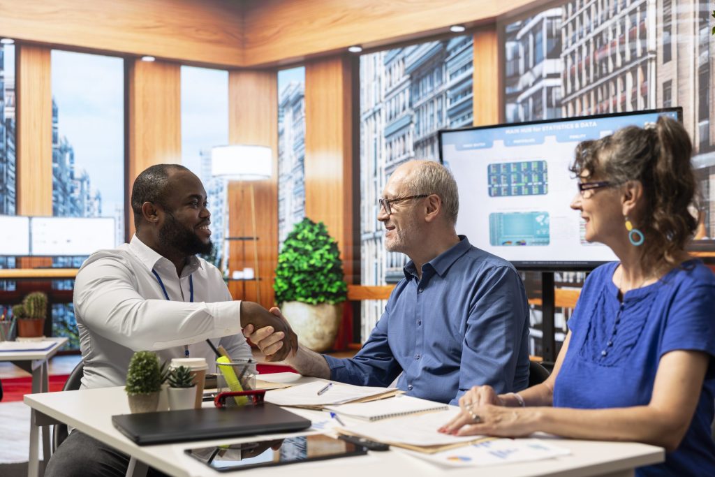 Three people sit at a desk in a modern office, two shaking hands and smiling while the third observes—perhaps finalizing an electronics recycling for insurance providers deal. Laptops, notebooks, and plants adorn the table, with city views through large windows.