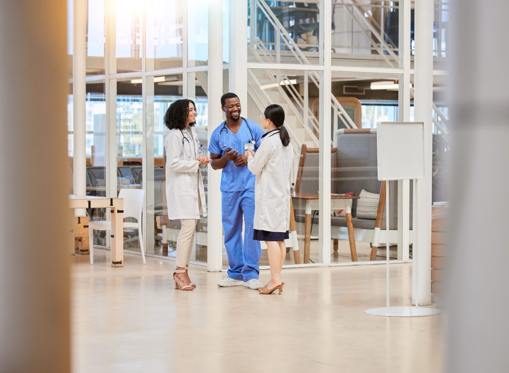 Three medical professionals, two women and one man, stand in a modern hospital atrium, smiling and talking together. The bright, open space highlights their commitment to HIPAA-compliant practices in every aspect of care.