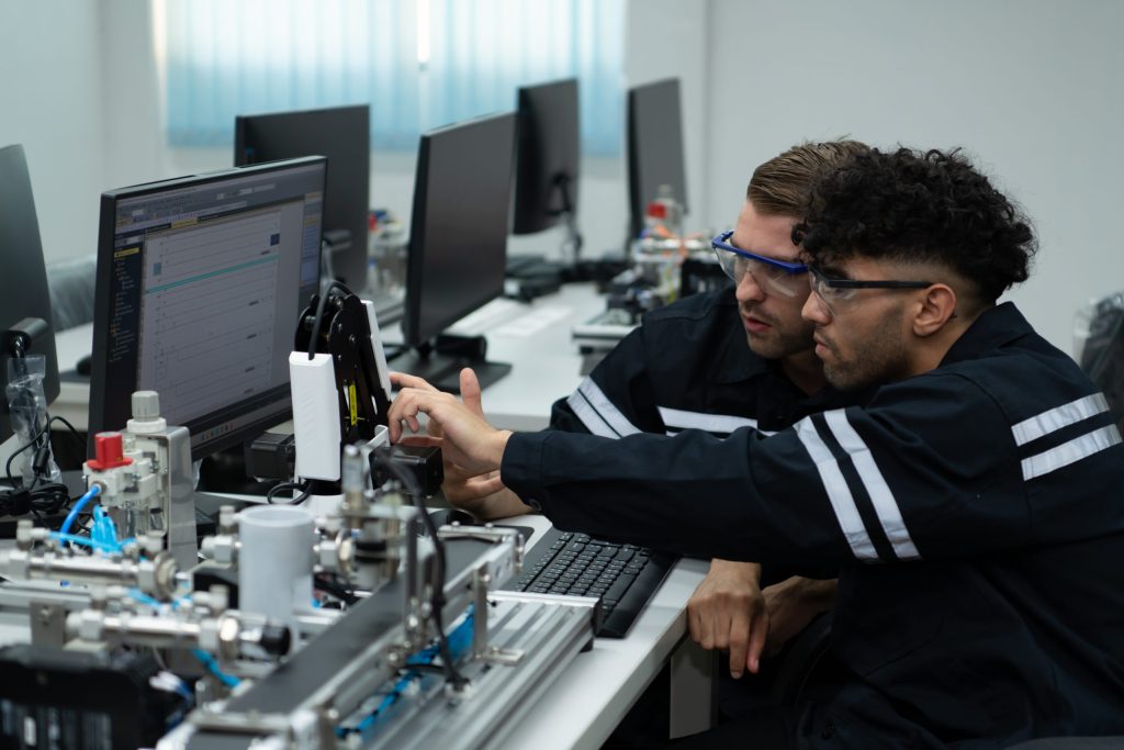 Two men wearing safety goggles and dark uniforms work together at a computer in a lab, analyzing electronics recycling for manufacturers data on the screen and handling technical equipment, surrounded by other monitors and machinery.