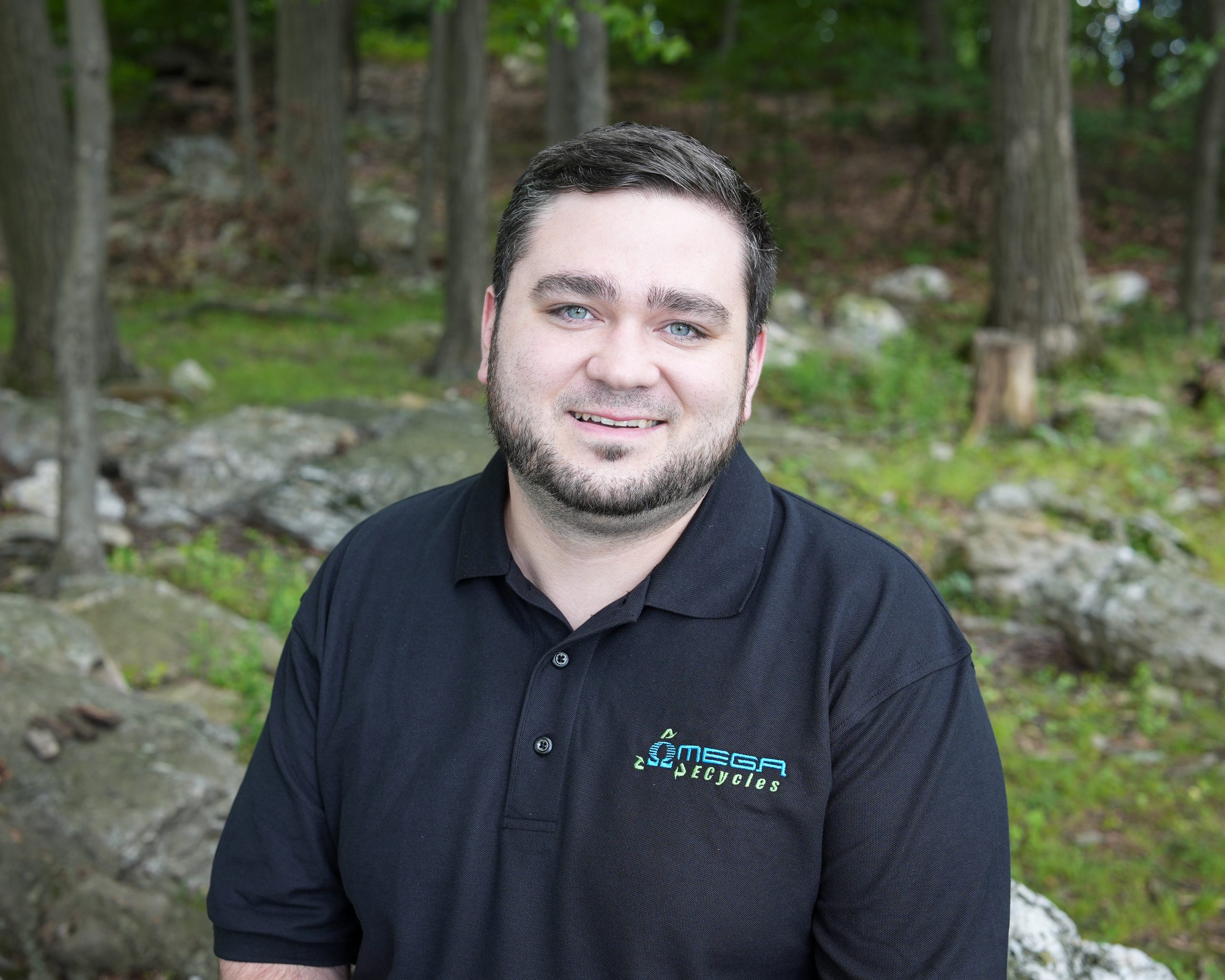 A man with short dark hair and a beard smiles at the camera, wearing a black polo shirt with the "Omega Recycles" logo, while standing in a wooded outdoor area. He is an Omega ECycles staff member.