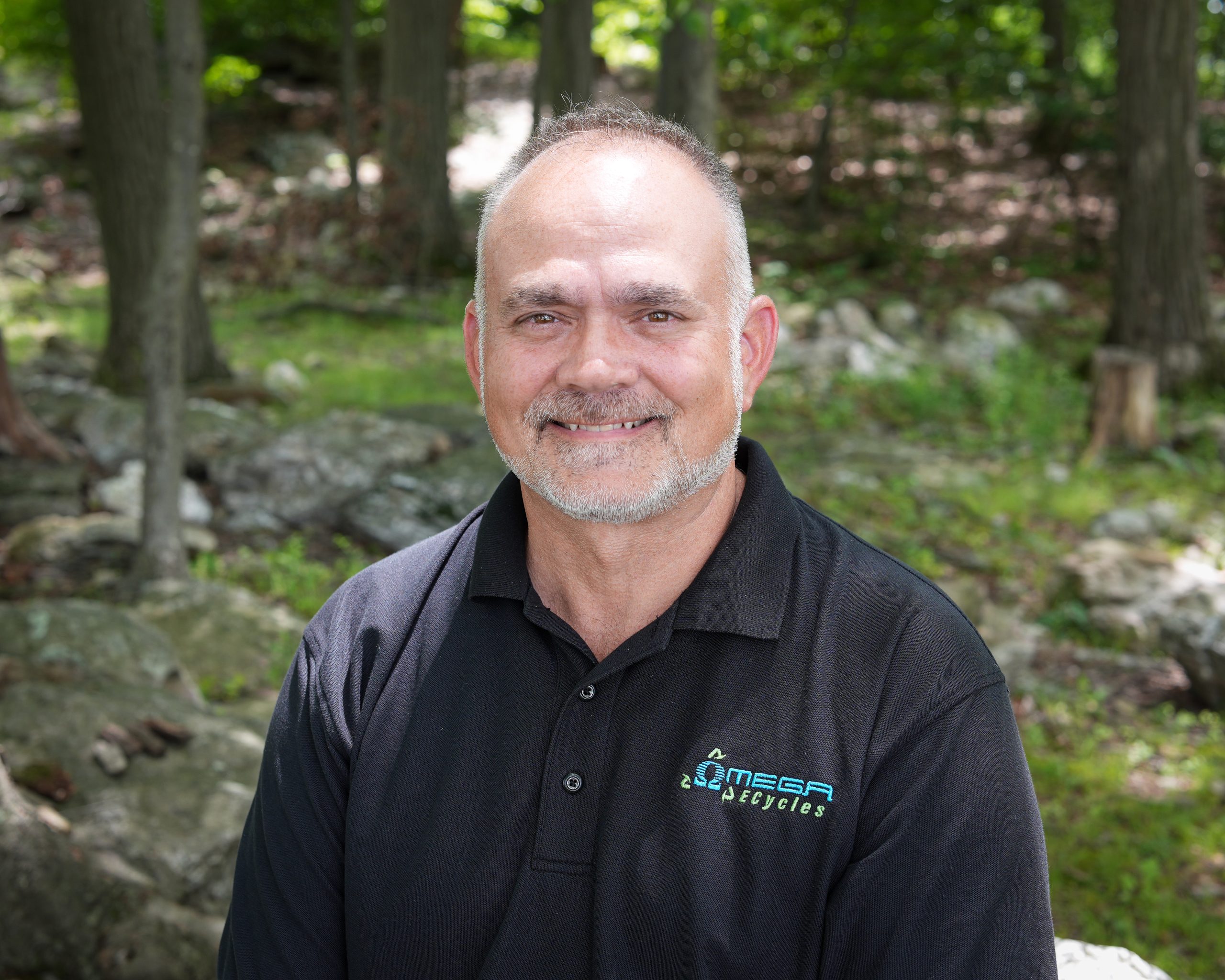Brent, an Omega ECycles staff member with a beard, smiles outdoors in a wooded area. He wears a black polo shirt with the "Omega Recycles" logo as sunlight filters through the trees in the background.
