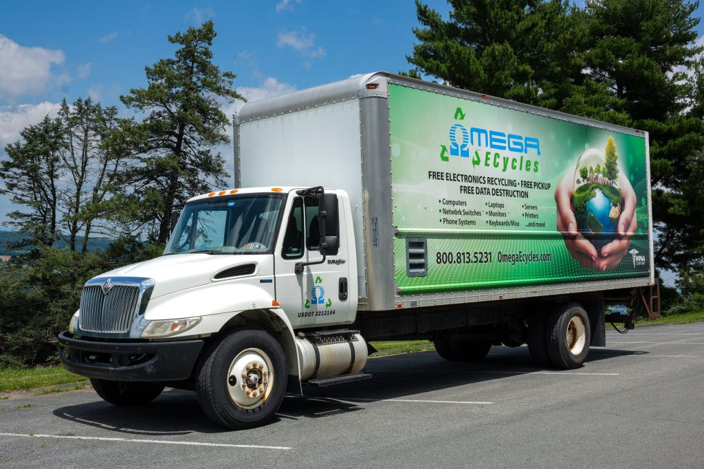A white box truck with "Omega E-cycles" branding is parked on pavement in Central PA. Advertising no cost electronics recycling, the truck features images of hands holding a green earth. Trees and a blue sky are in the background.