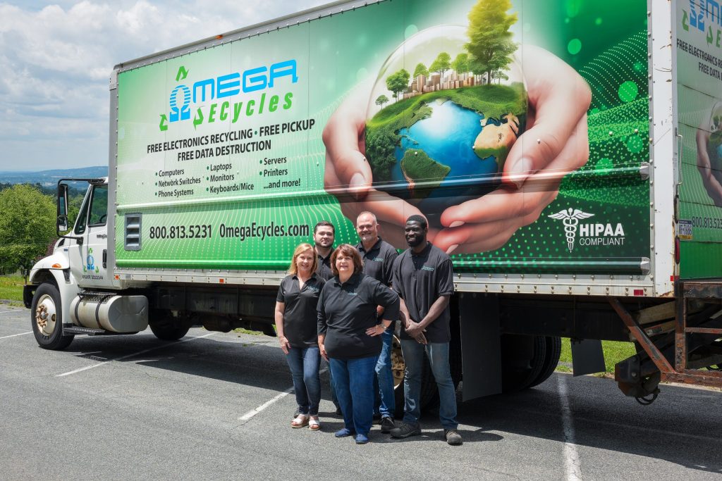 Five people stand smiling in front of a MEGA Recycles truck with a graphic of the Earth and recycling imagery. The Omega ECycles team, appearing as staff members, promotes electronics recycling and free pickup services.