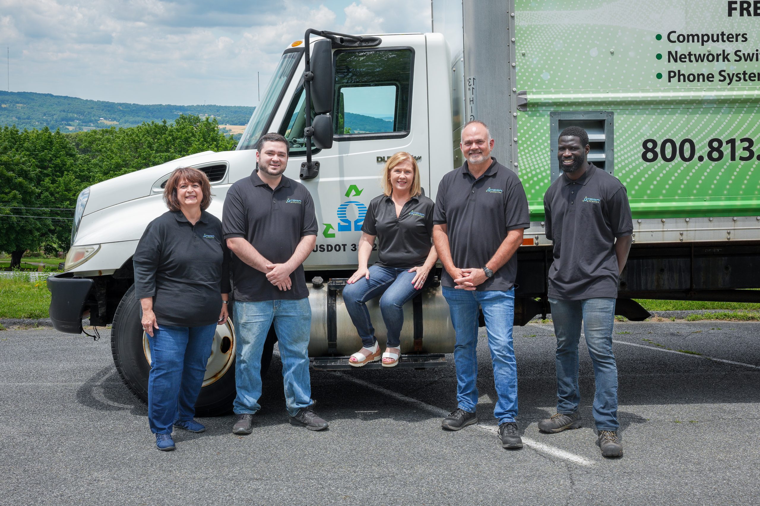 Five smiling members of the Omega ECycles team in matching black shirts pose in front of a white delivery truck with a green and blue logo. One woman sits on the truck step while others stand, framed by trees and hills in the background.