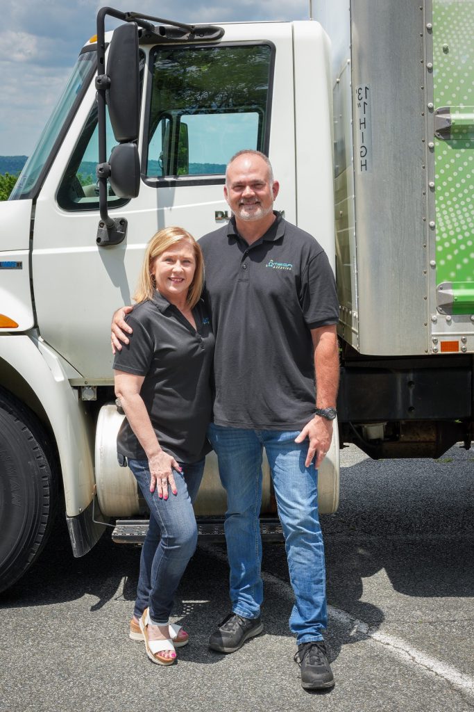 Reesy and Brent from Omega ECycles stand smiling with arms around each other in front of a white truck, both wearing matching black company polo shirts and jeans on a sunny day.