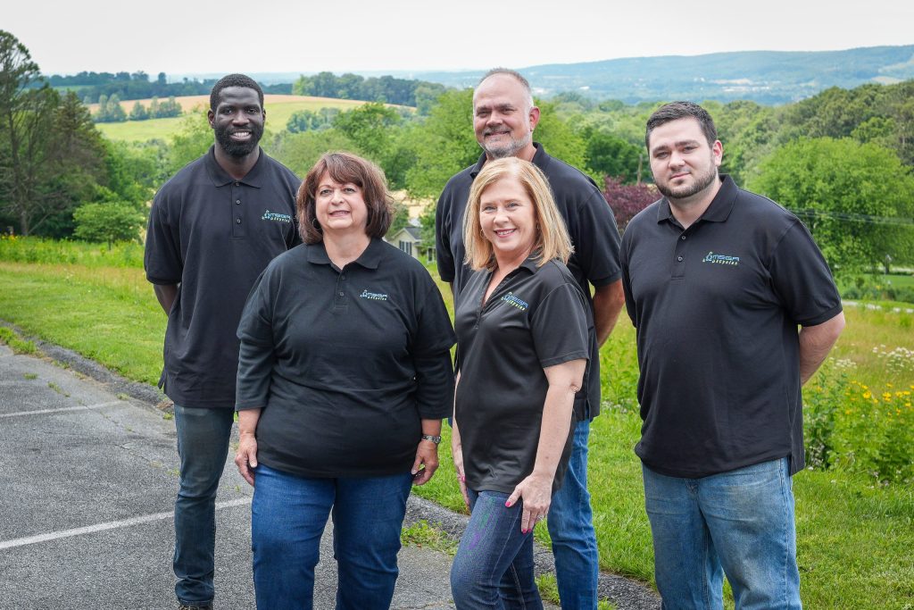 Five adults from the Omega ECycles team stand outdoors on a paved path with green hills and trees in the background, all wearing black collared shirts and smiling at the camera.
