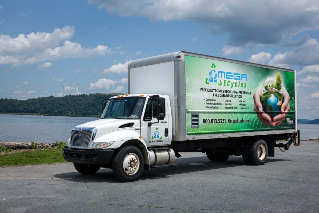 A white Omega Ecycles recycling truck, promoting Free Electronics Pickup, is parked by a body of water with hills and wind turbines in the background. The truck features a green advertisement for free electronics recycling services.