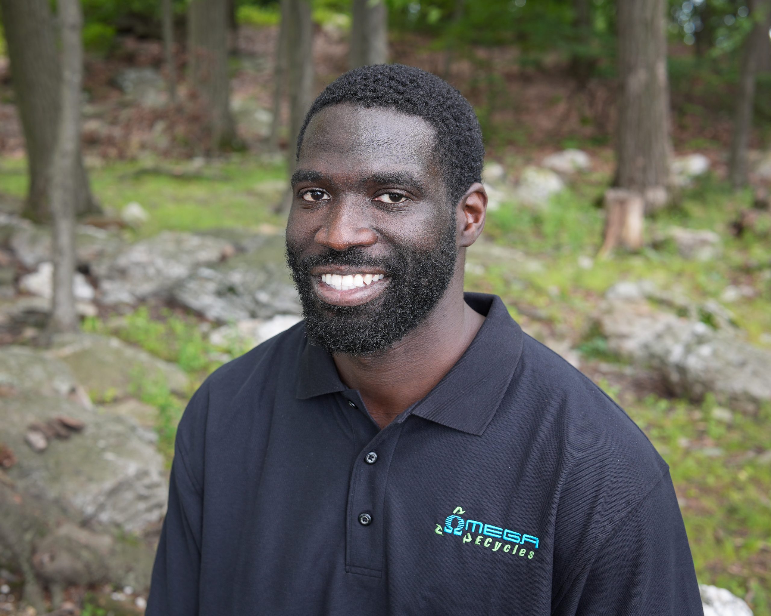 A man with a beard and short curly hair smiles at the camera. Jere, an Omega ECycles staff member, is outdoors in a black Omega Cycles polo shirt, surrounded by green grass, rocks, and trees.
