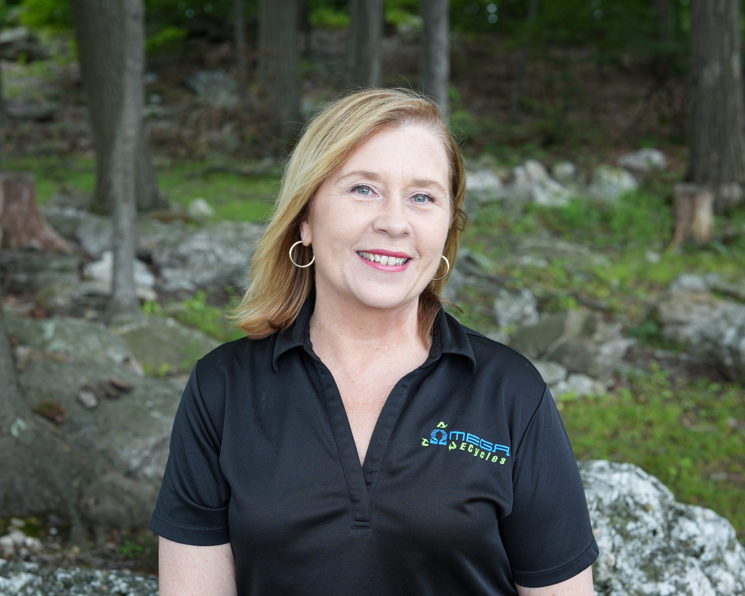 A woman with shoulder-length blonde hair, wearing hoop earrings and a black polo shirt with an embroidered "OMEGA" logo, stands smiling outdoors. Meet Reesy, Omega ECycles founder, surrounded by a forested area with rocks and trees in the background.