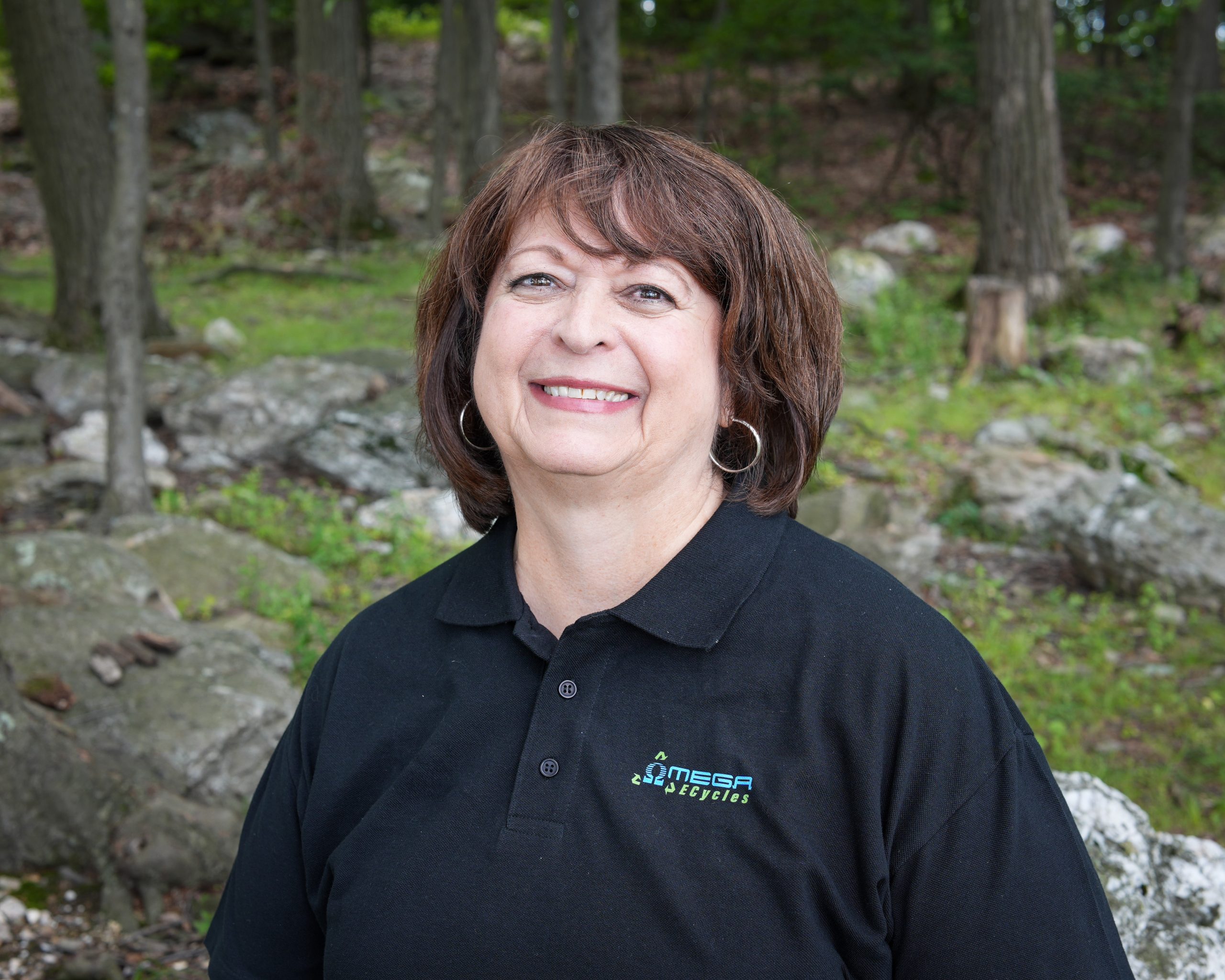 Wendy, an Omega ECycles staff member with short brown hair and hoop earrings, smiles outdoors in a wooded area with rocks and trees, wearing a black "OMEGA" logo polo shirt.