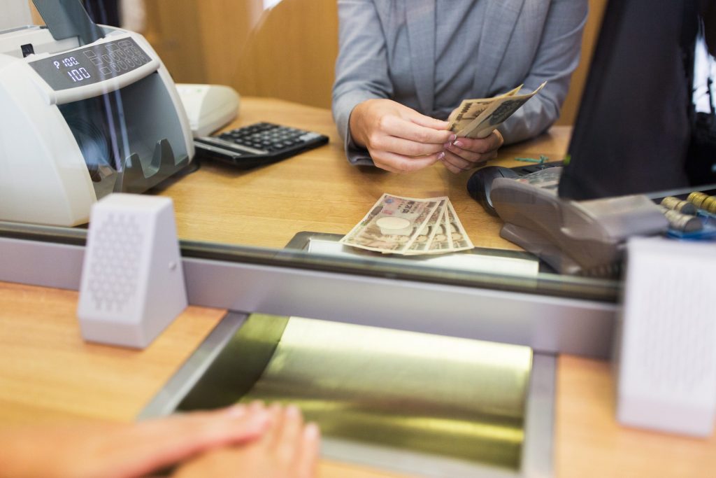 A bank teller behind a glass partition counts cash bills at one of the leading banks, while a customer waits on the other side with hands resting on the counter. A calculator and currency counting machine are visible on the desk.