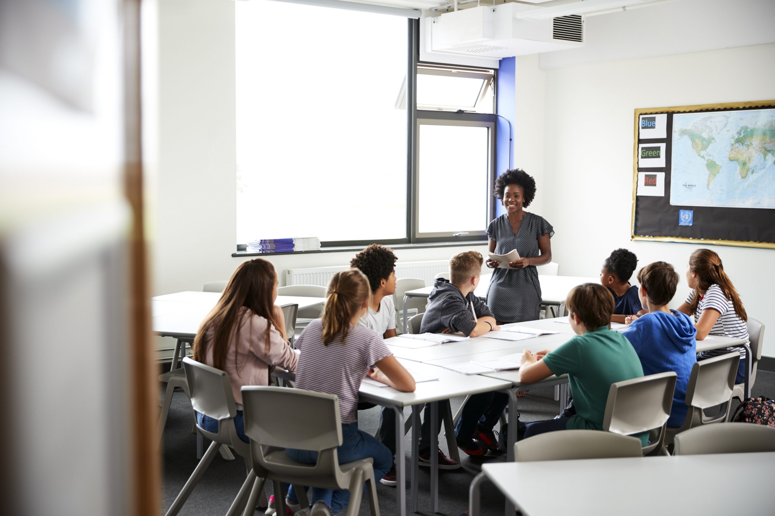 A teacher stands at the front of a classroom, smiling and speaking to a group of students seated around tables. The classroom, like many schools, has large windows, maps on the wall, and natural light streaming in.