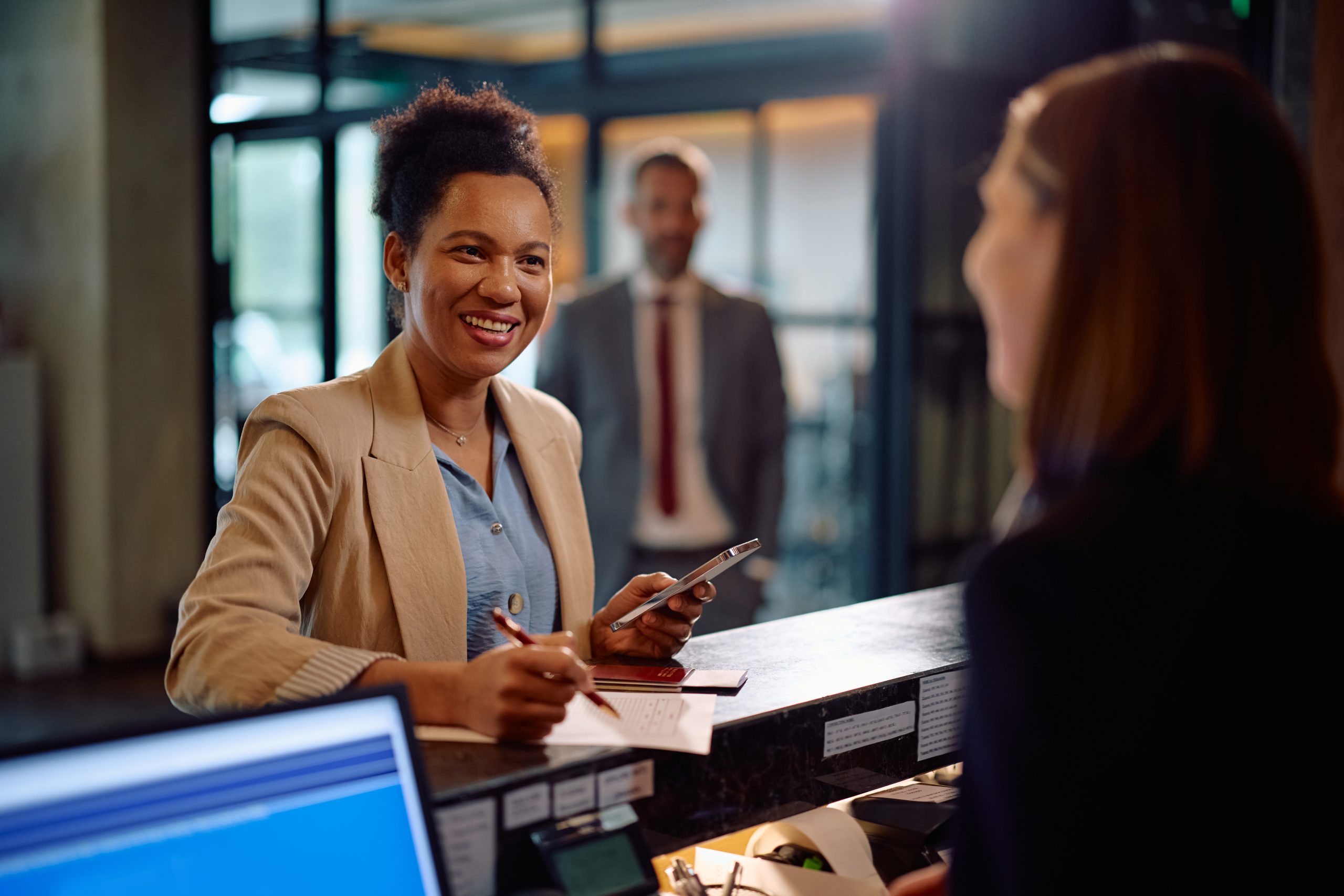 A woman smiles while checking in at a reception desk, holding a phone and a pen. She speaks with a receptionist about Omega ECycles' electronics recycling for banks as a man in a suit waits in the background of the office lobby.