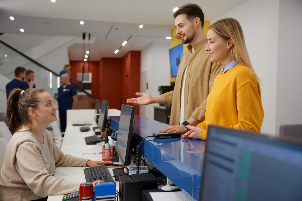 A smiling woman and man stand at a reception desk, interacting with a seated receptionist who is also smiling. Omega ECycles brochures about electronics recycling for banks are visible among the computer monitors and office equipment in the modern office.