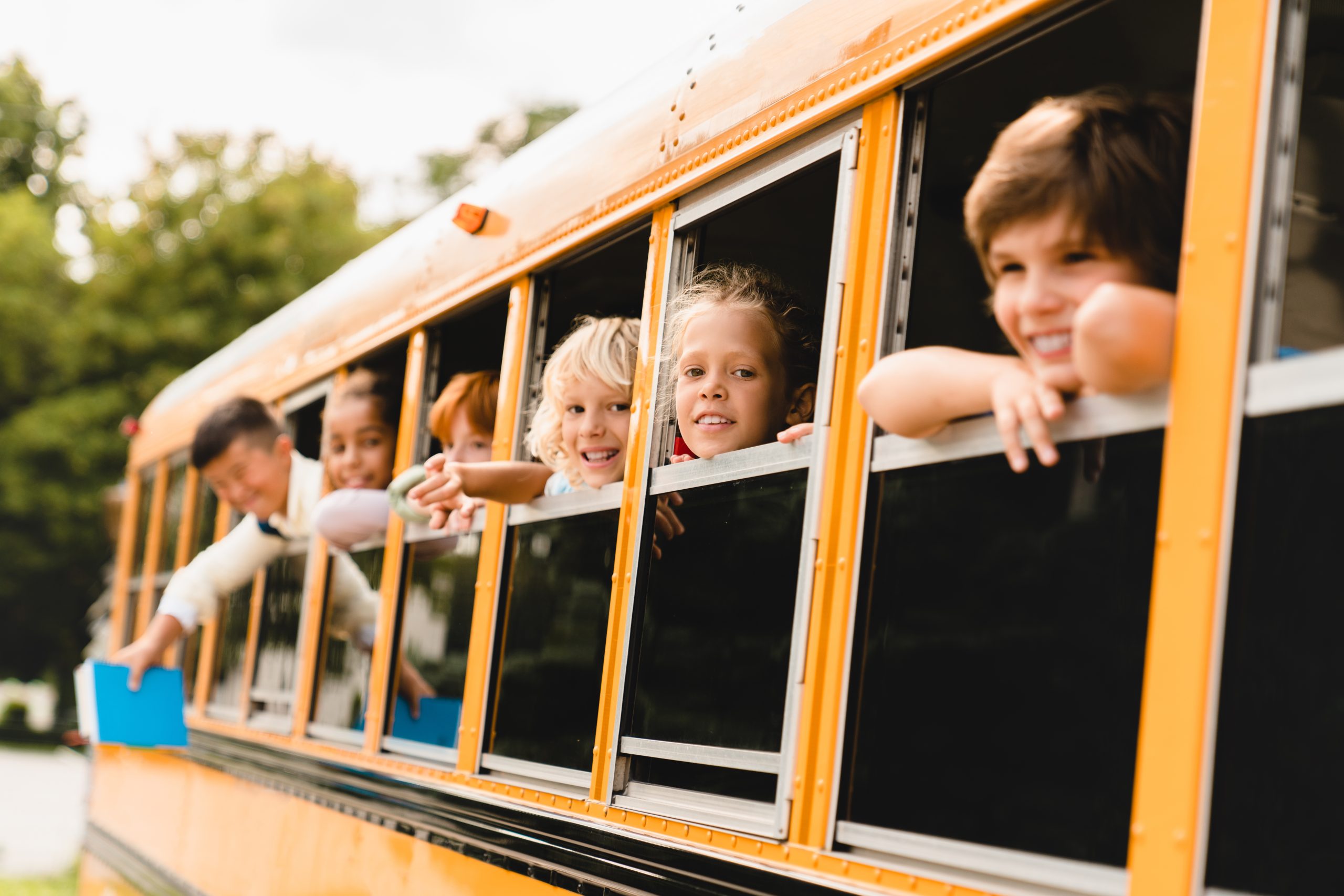 A group of smiling children lean out of the windows of a yellow school bus during a Stuff-A-Bus event, looking excited and happy as they collect school supplies. Trees are visible in the blurred background.