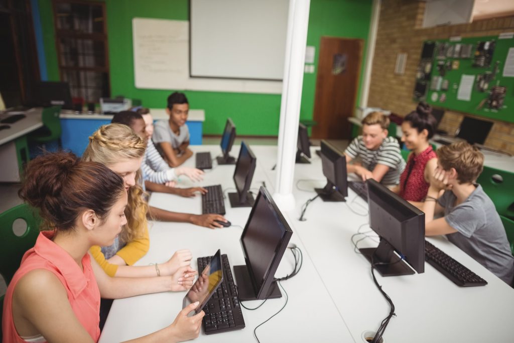 A group of students sits at desks in a computer lab, working at desktop computers and collaborating in small groups. The bright, modern setting with green walls promotes learning and highlights Omega ECycles’ commitment to outdated electronics recycling.