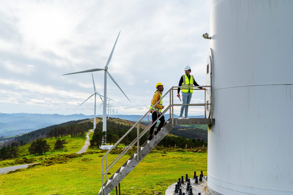 Two workers wearing safety helmets and vests stand on stairs leading to a wind turbine operated by an energy company, with wind turbines and green hills visible in the background under a cloudy sky.