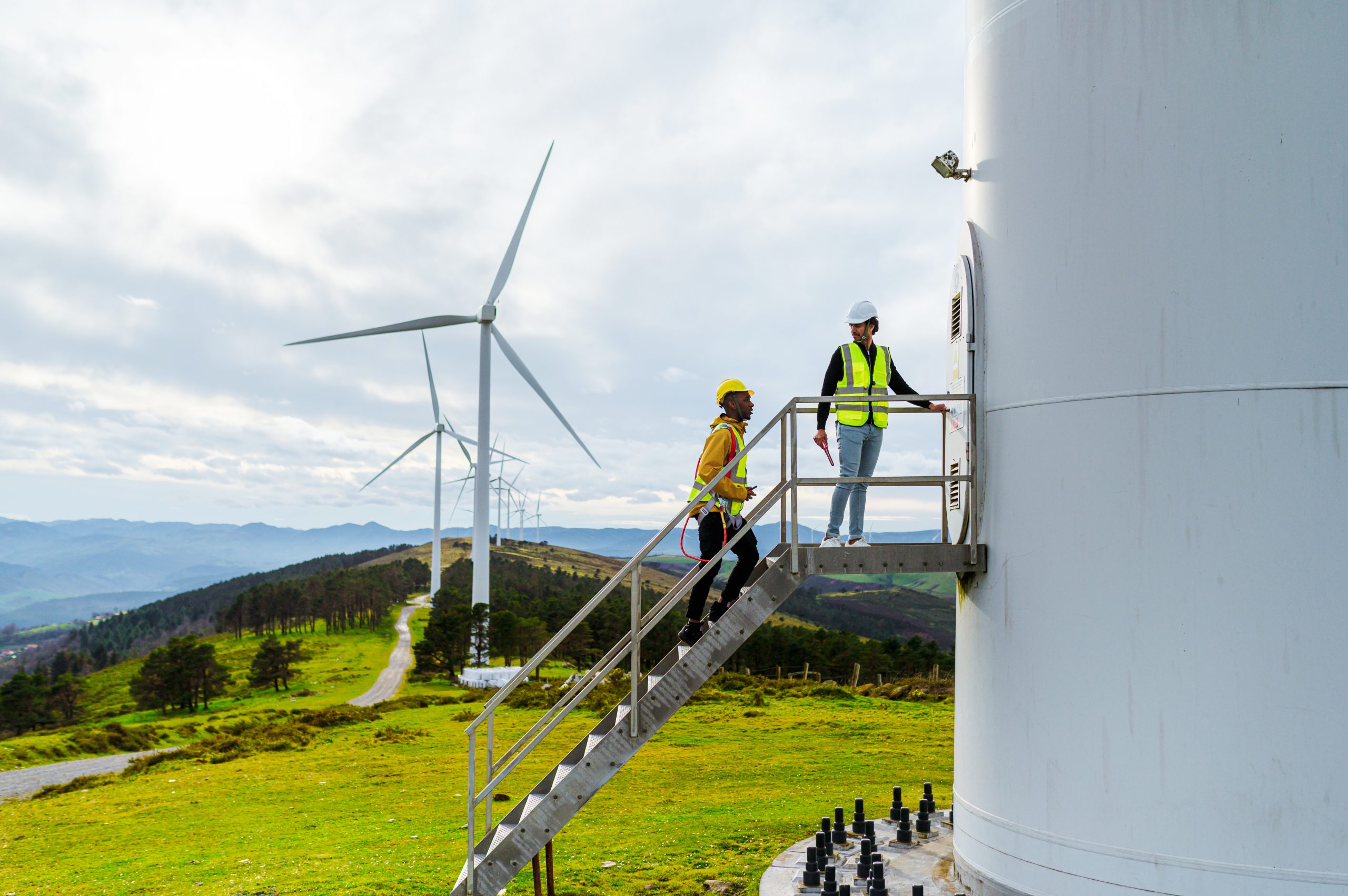 Two workers wearing safety helmets and vests stand on stairs leading to a wind turbine operated by an energy company, with wind turbines and green hills visible in the background under a cloudy sky.