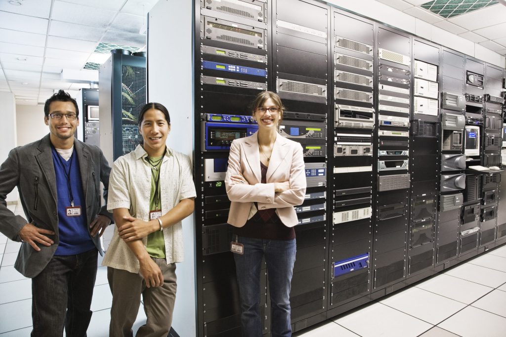 Three people stand in front of tall racks of computer servers in a brightly lit server room, smiling at the camera. Representing IT companies, they wear ID badges and business casual clothing, ready to discuss free electronics recycling solutions.