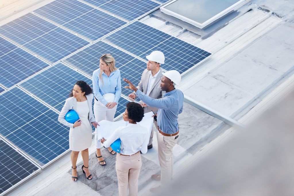 A group of five professionals from an energy company, some wearing hard hats and holding blueprints or helmets, stand on a rooftop covered with solar panels, discussing the project.