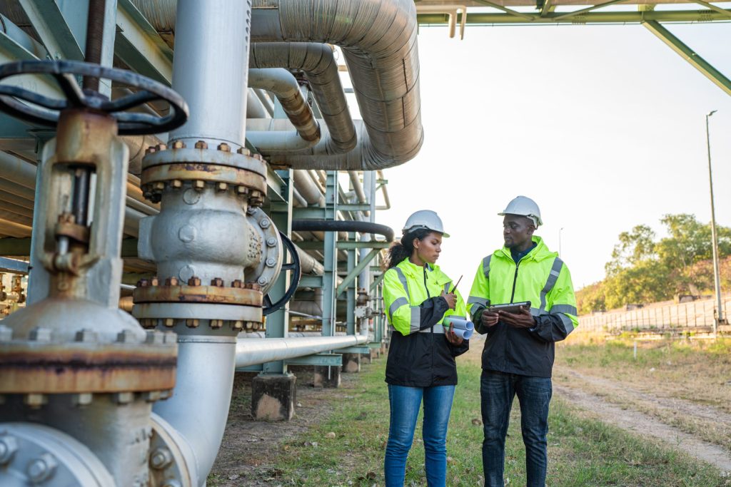 Two workers in safety jackets and helmets stand outdoors by large industrial pipes at an energy company, discussing something while holding a tablet and clipboard.