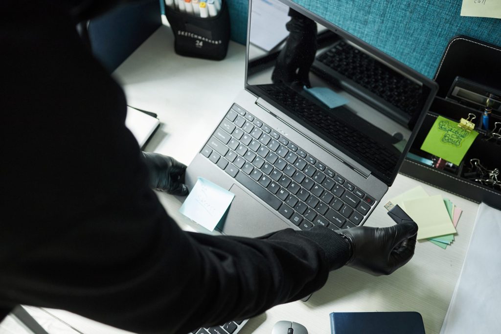 A person wearing black gloves places a sticky note on a laptop keyboard, possibly hinting at secretive laptop recycling activity. Various office supplies and documents are scattered on the desk, adding to the scene’s intrigue.