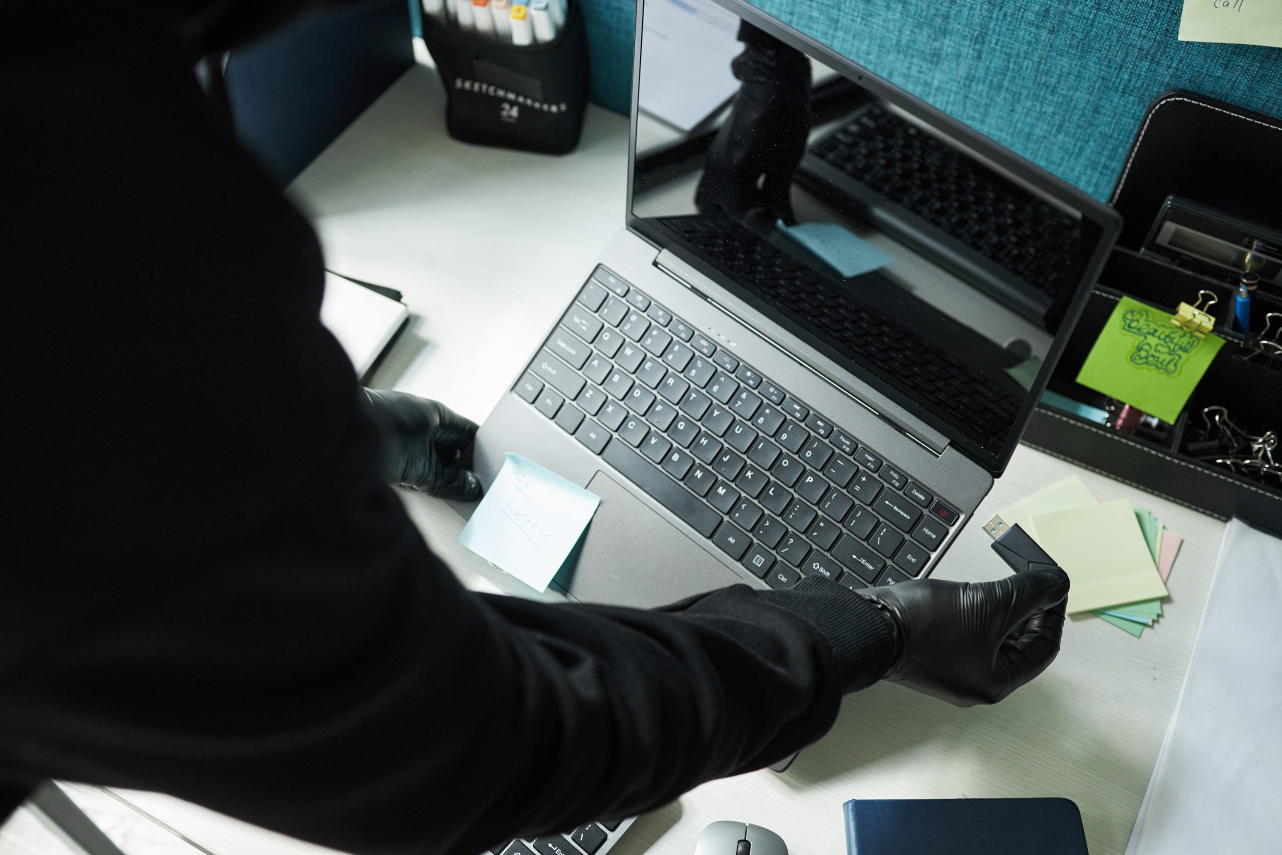 A person wearing black gloves places a sticky note on a laptop keyboard, possibly hinting at secretive laptop recycling activity. Various office supplies and documents are scattered on the desk, adding to the scene’s intrigue.
