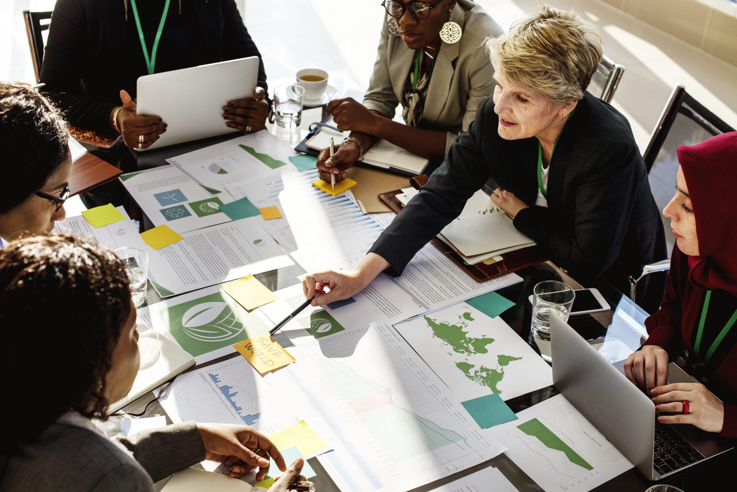 A diverse group of professionals gathers around a conference table covered with charts, graphs, and documents, collaborating and discussing data to develop a sustainable business strategy during a meeting.