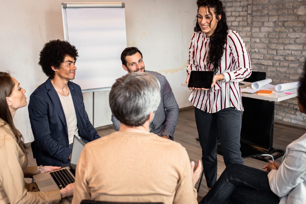 A woman stands smiling with a tablet, presenting to four seated colleagues in a casual office setting with a flip chart and laptops, as she trains employees to properly retire devices.