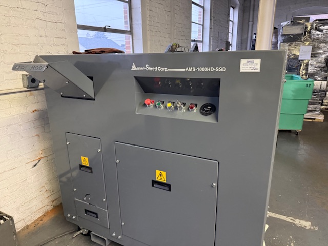 A large, gray Ameri-Shred Corp. AMS-1000HD-SSD industrial shredder stands in a warehouse near a brick wall, with various buttons and warning labels visible on its front panel.