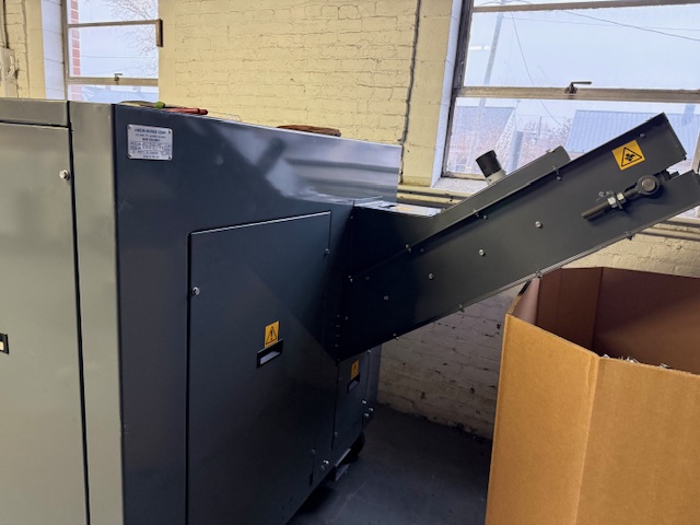 An Ameri-Shred Corp. AMS-1000HD-SSD industrial machine with a slanted conveyor chute sits next to a cardboard box in a warehouse with brick walls and tall windows. Safety warning labels are visible on the machine.