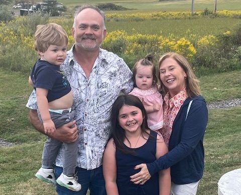 A smiling family of five, including Omega ECycles founder Reesy Neff, poses outdoors in a grassy field. The man holds a young boy, the woman holds a baby girl, and an older girl stands in front. Yellow flowers brighten the background.