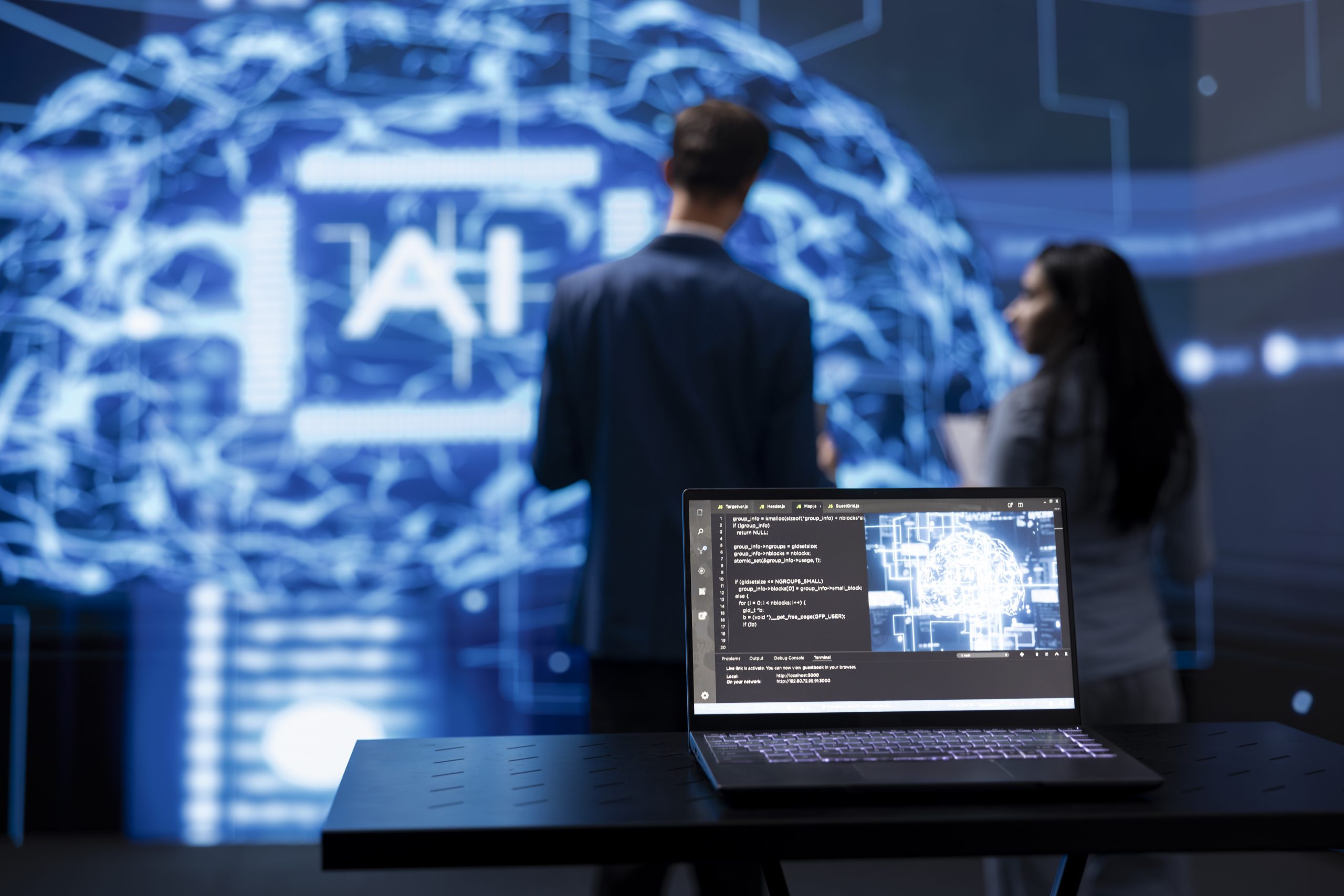 A laptop displaying code sits on a table in the foreground, while two blurred people stand before a large digital screen showing a glowing AI brain—highlighting the risks of improper device retirement in tech-driven environments.