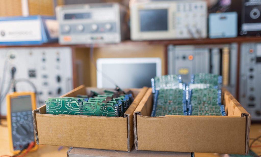 Two cardboard boxes filled with green circuit boards sit on a workbench at an electronics recycling facility, with various electronic testing equipment and instruments blurred in the background.