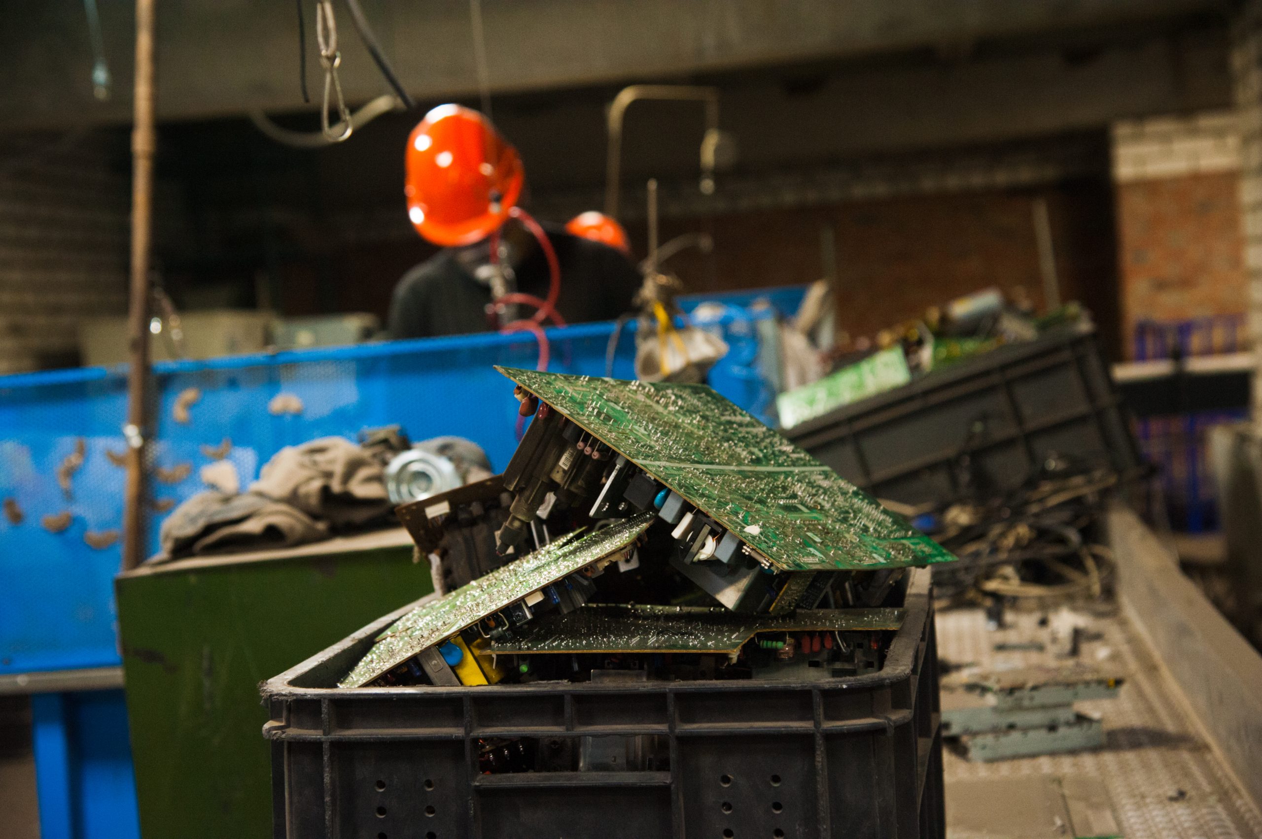 A black crate filled with discarded electronic circuit boards, destined for e-recycling, sits in the foreground of a factory setting, while workers in orange helmets work in the blurred background.