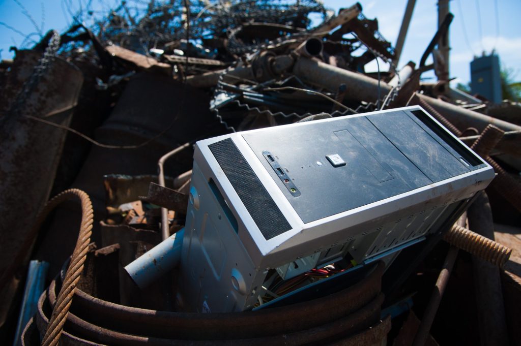 A discarded computer tower lies among a pile of metal debris and rusty cables outdoors, awaiting electronic recycling under a bright blue sky.