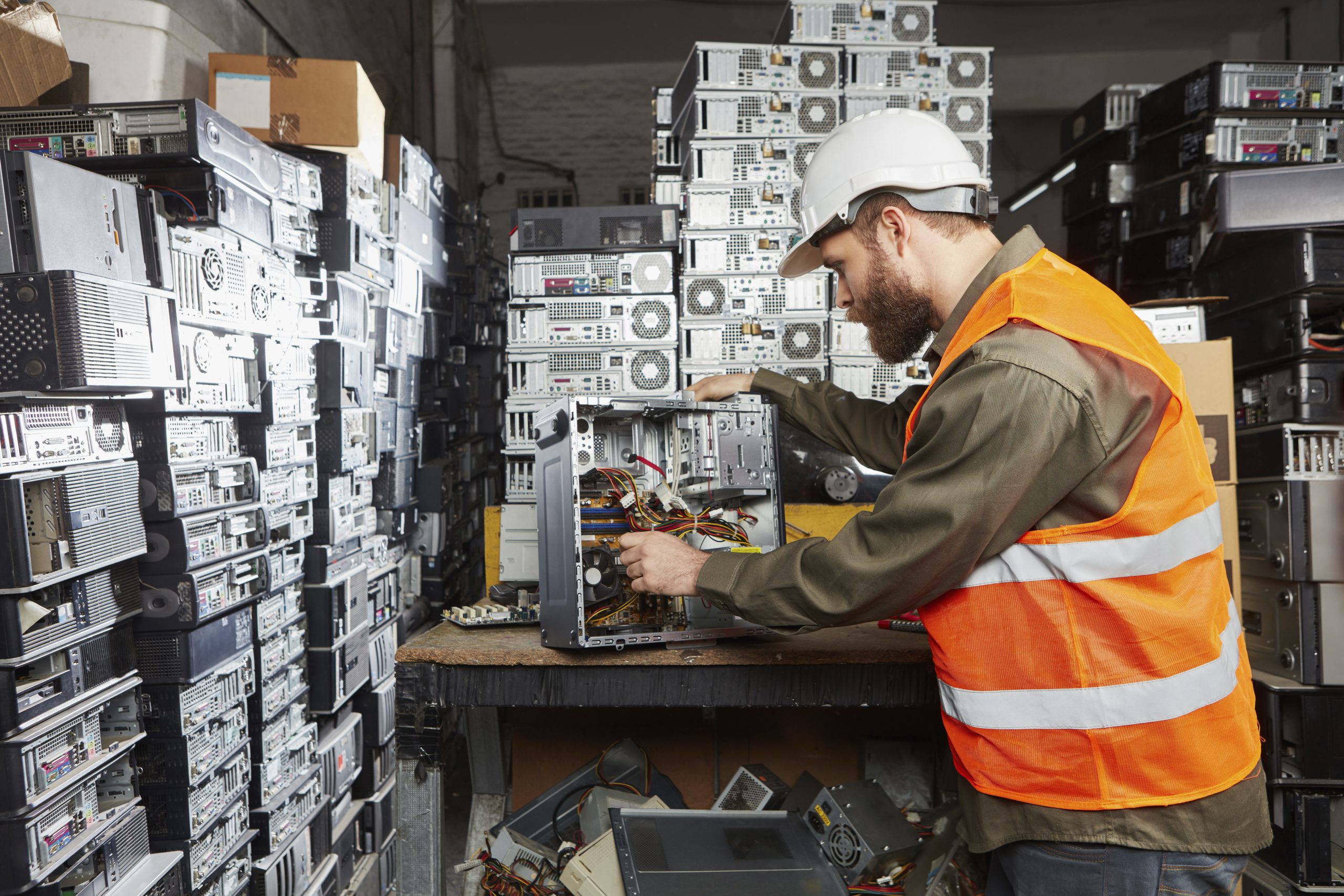 A man in a white hard hat and orange safety vest repairs a desktop computer at a workstation, surrounded by large stacks of used towers in a computer recycling facility.
